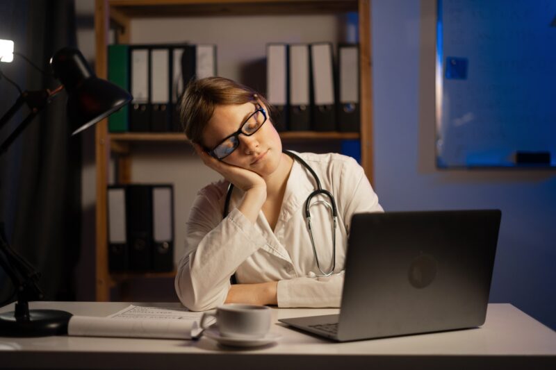 Medizinstudentin mit Stethoskop sitzt müde vor einem Laptop im Büro, stützt den Kopf auf die Hand – symbolisiert Erschöpfung, lange Arbeitszeiten oder Belastung im medizinischen Beruf.
